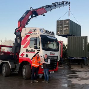 Craig from CTS presenting Simon from Bayliss Haulage with his Transport Manager CPC certificate. They are stood in front of a Bayliss Haulage HIAB lifting a container.