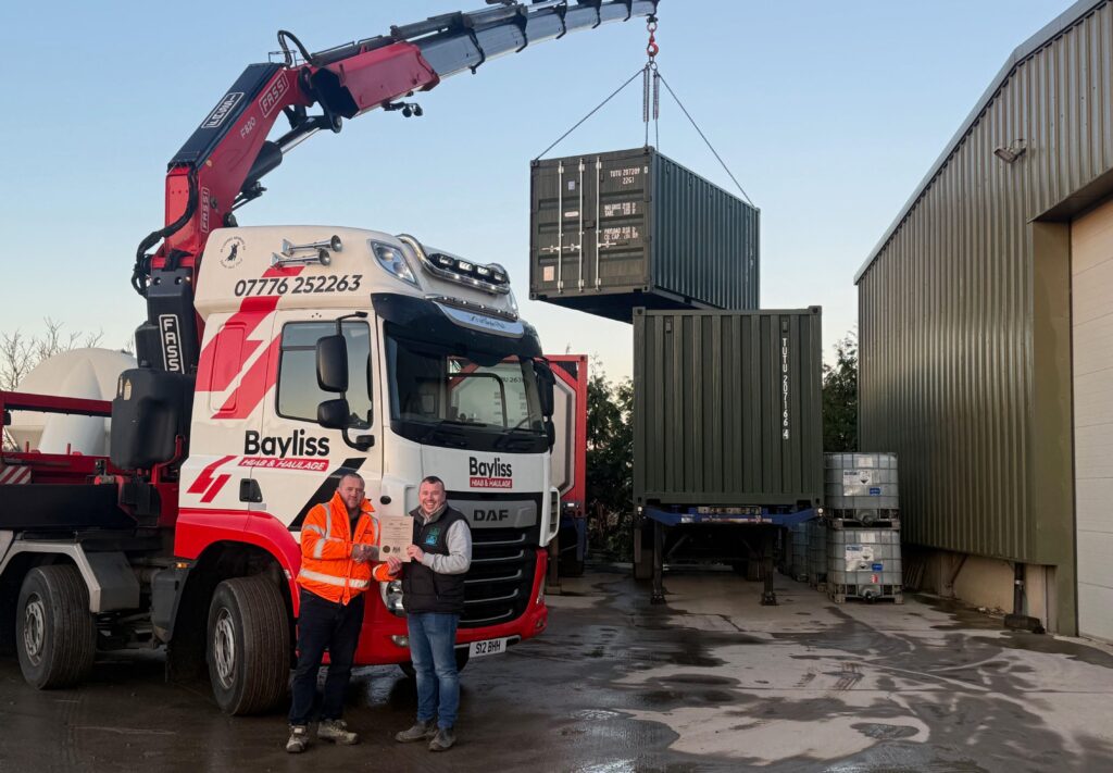 Craig from CTS presenting Simon from Bayliss Haulage with his Transport Manager CPC certificate. They are stood in front of a Bayliss Haulage HIAB lifting a container.